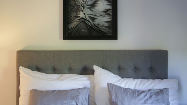 Un letto con testiera grigia rivestita in tessuto, due cuscini bianchi e un quadro appeso sopra.