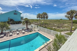 Un espace piscine avec vue sur la plage, un bâtiment doté d’un balcon et une terrasse clôturée avec des chaises longues.
