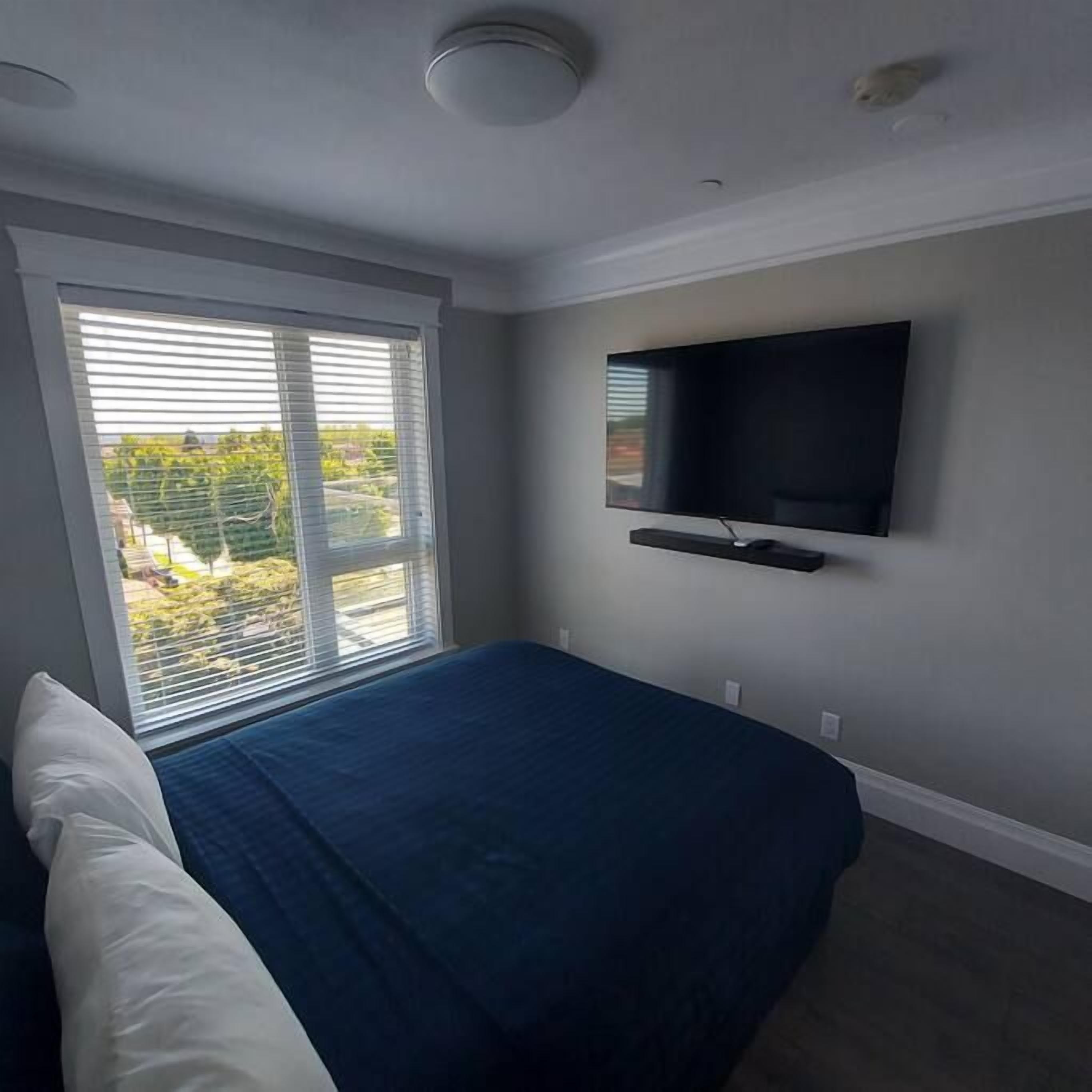 A bedroom with a bed, a window with blinds, a mounted TV, and a ceiling light.