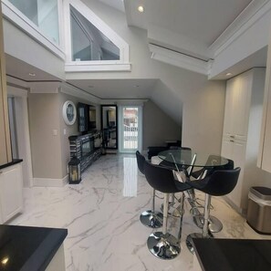 A modern kitchen with a marble countertop, a dining area with a glass table and black chairs, and a view of the backyard through a large window.