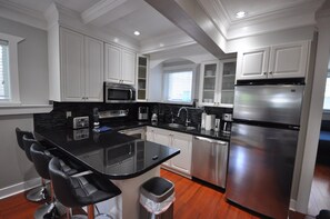 A modern kitchen with white cabinetry, stainless steel appliances, a black countertop, and a central island with bar stools. - New Luxurious 3 Bedroom Kingsway Castle Suite (Burnaby)