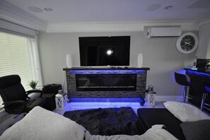 A modern living room with a flat-screen TV mounted on a wall, a fireplace with a blue light, a comfortable seating area with a white furry rug, and a small side table with a plant. - Beautiful Luxurious 3 Bed Townhouse (Vancouver)