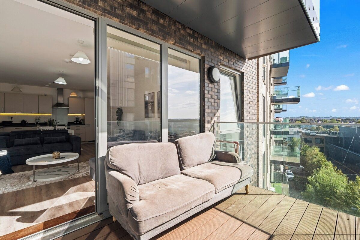 A balcony with a sofa and a dining table, a kitchen in the background, and a view of the city.