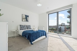 A bedroom with a large window, a bed with a blue blanket, a white radiator, and a small bedside table.
