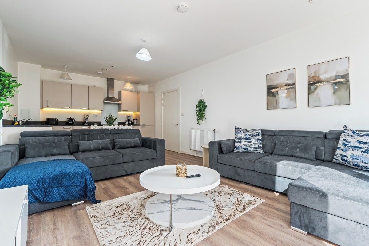 A modern living room with grey sofas, a white coffee table, and a kitchen in the background.