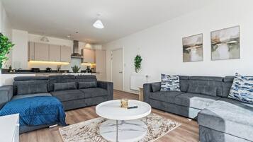 A modern living room with grey sofas, a white coffee table, and a kitchen in the background.