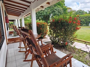 A covered outdoor seating area with wooden chairs and tables, surrounded by lush greenery and flowering plants.