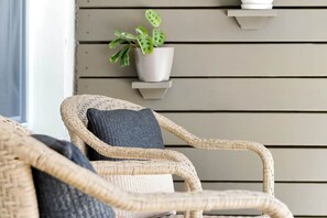 Une chaise en osier avec un coussin foncé, une plante en pot et une étagère fixée au mur, le tout placé devant un mur lambrissé.