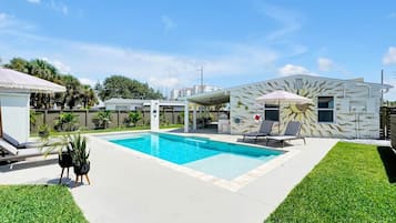 Ein Swimmingpool mit Liegestühlen und Sonnenschirmen, umgeben von einem gepflegten Rasen und einem Gebäude mit markanter Fassade.