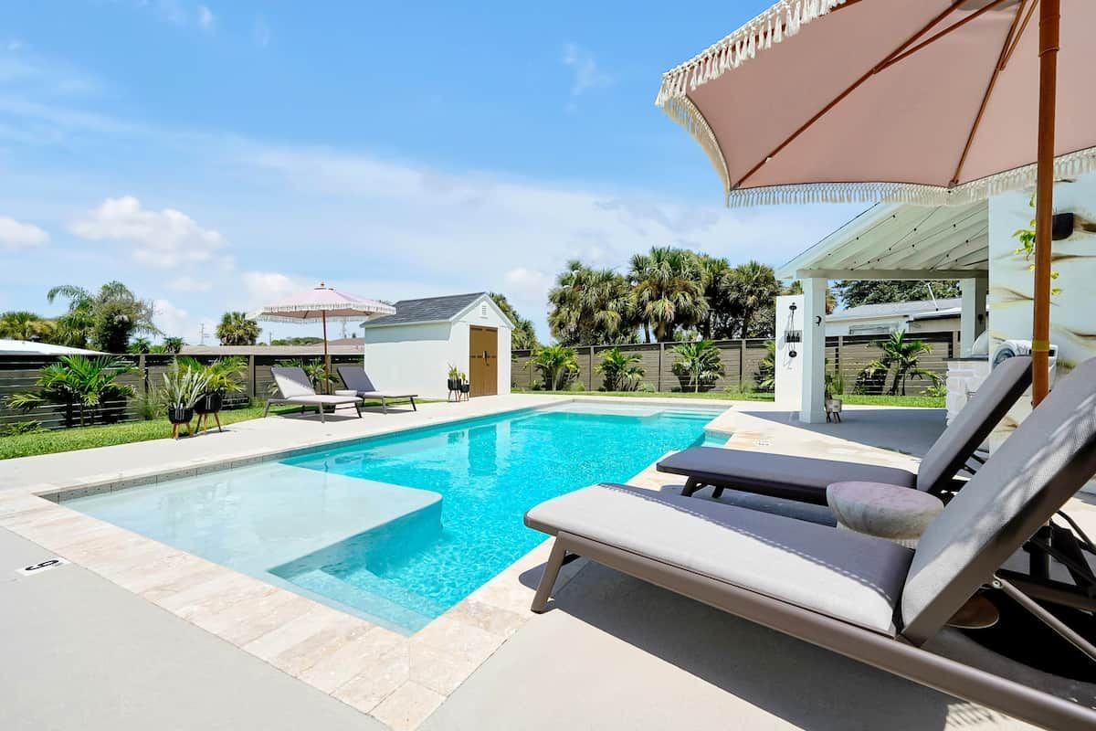 Ein Pool mit Liegestühlen und Sonnenschirmen, umgeben von einer Umzäunung und Palmen.