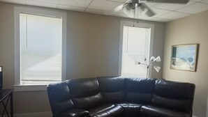 Una sala de estar con un sofá modular, dos ventanas con persianas, un ventilador de techo y un cuadro en la pared.