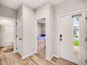 Un couloir doté d’un plancher en bois, de murs blancs et d’une porte donnant sur une chambre à coucher, où se trouvent un lit et un ventilateur de plafond.