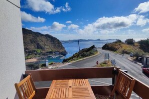 A balcony with a wooden table and chairs overlooking a coastal road with a view of the sea and hills. - Machico Terrace (Machico)