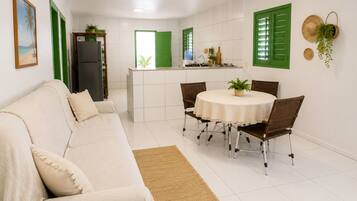 A modern living room with a sofa, a dining table with chairs, a kitchen area with a refrigerator, and a green door.
