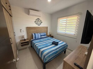 A bedroom with a bed, bedside tables, a TV, and a window with blinds. - Enjoy swimming pools, a rooftop area with a Jacuzzi, and a BBQ grill. (Mazatlán)