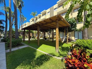 A covered outdoor area with a wooden pergola, surrounded by palm trees and well-maintained grass. - Enjoy swimming pools, a rooftop area with a Jacuzzi, and a BBQ grill. (Mazatlán)