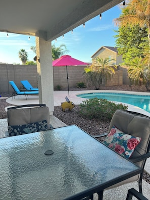 An outdoor patio area with a glass-top table, two lounge chairs, a decorative pig statue, and a swimming pool. - Pet Friendly-Priv. Pool-King Suite-Golf Nearby (San Tan Valley)