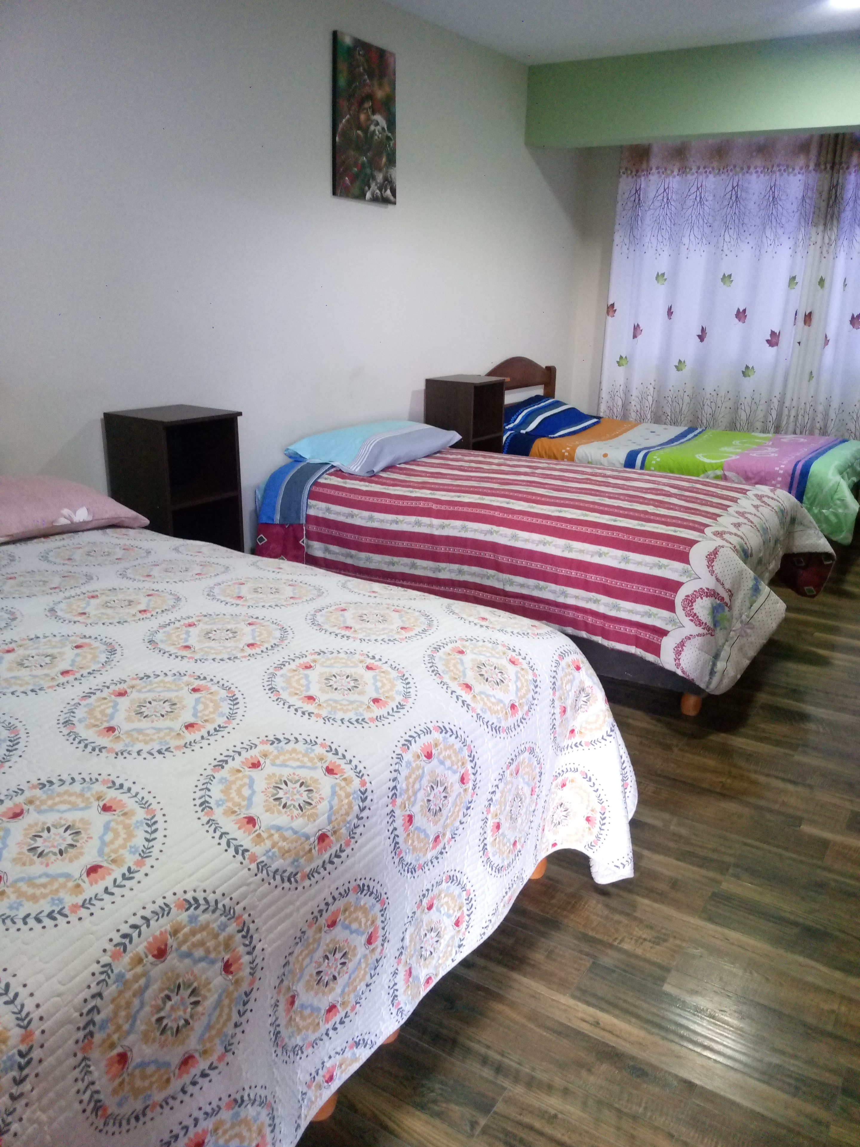 Una habitación con tres camas, cada una con una colcha de diseño diferente. Hay dos mesitas de noche y una repisa en la pared.