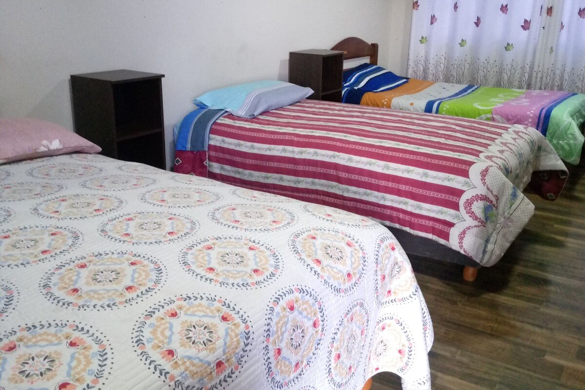 A room with three beds, each with a different patterned bedspread. There are two nightstands and a wall-mounted shelf.