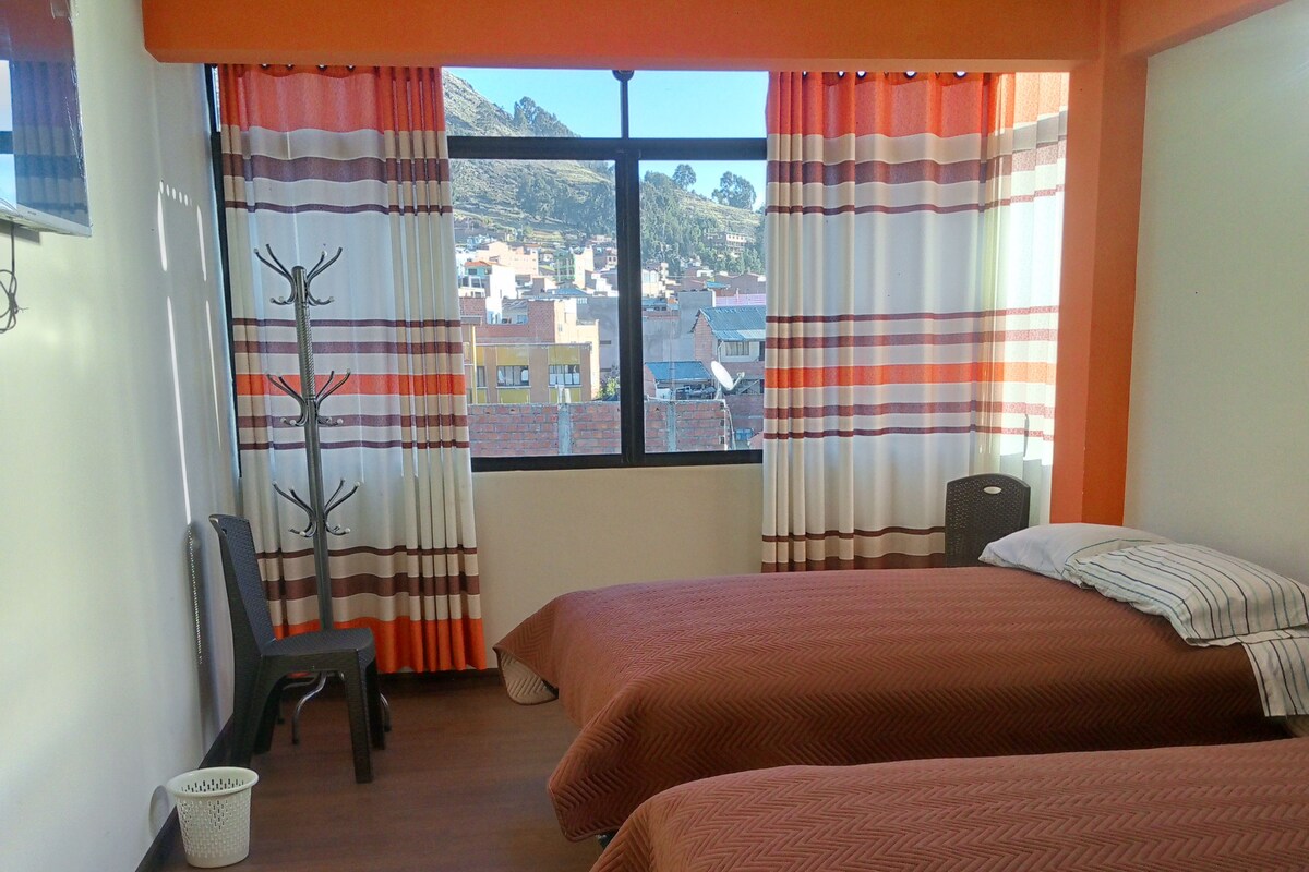 A hotel room with two beds, a chair, a coat rack, and a view of the cityscape through the window.
