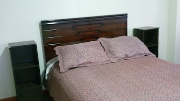 A bedroom with a wooden bed, a patterned bedspread, a wall-mounted artwork, and a small shelf.