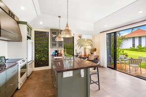 Une cuisine moderne dotée d’un îlot central, de luminaires suspendus et offrant une vue sur un jardin tropical.