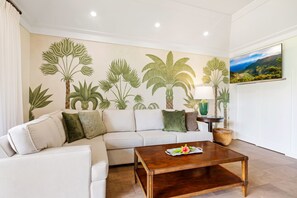 A living room with a white sofa, a wooden coffee table, and a wall mural of tropical plants. - Kiahuna 150 – Fully Remodeled, A/C, Near Beach (Koloa)