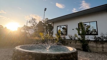 En rund stenfountain med vandstråler, omgivet af grus, foran en moderne bygning med store vinduer.