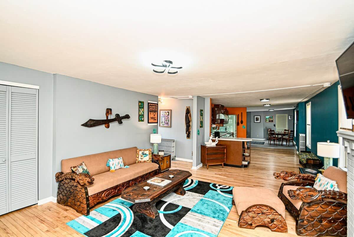 A living room with a sofa, coffee table, and patterned rug. There is a kitchen area in the background with a refrigerator and a dining table.