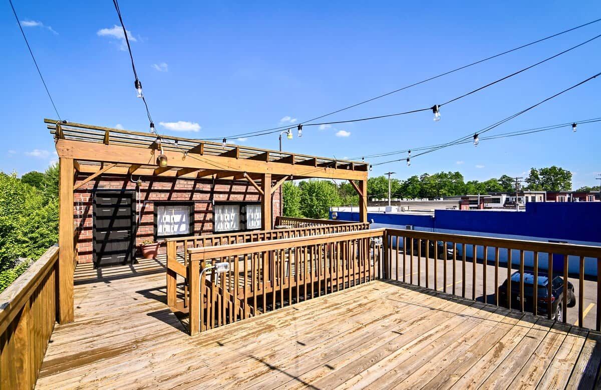 A wooden deck with a pergola structure, a small enclosed area with a door, and a view of a parking lot and buildings in the background.
