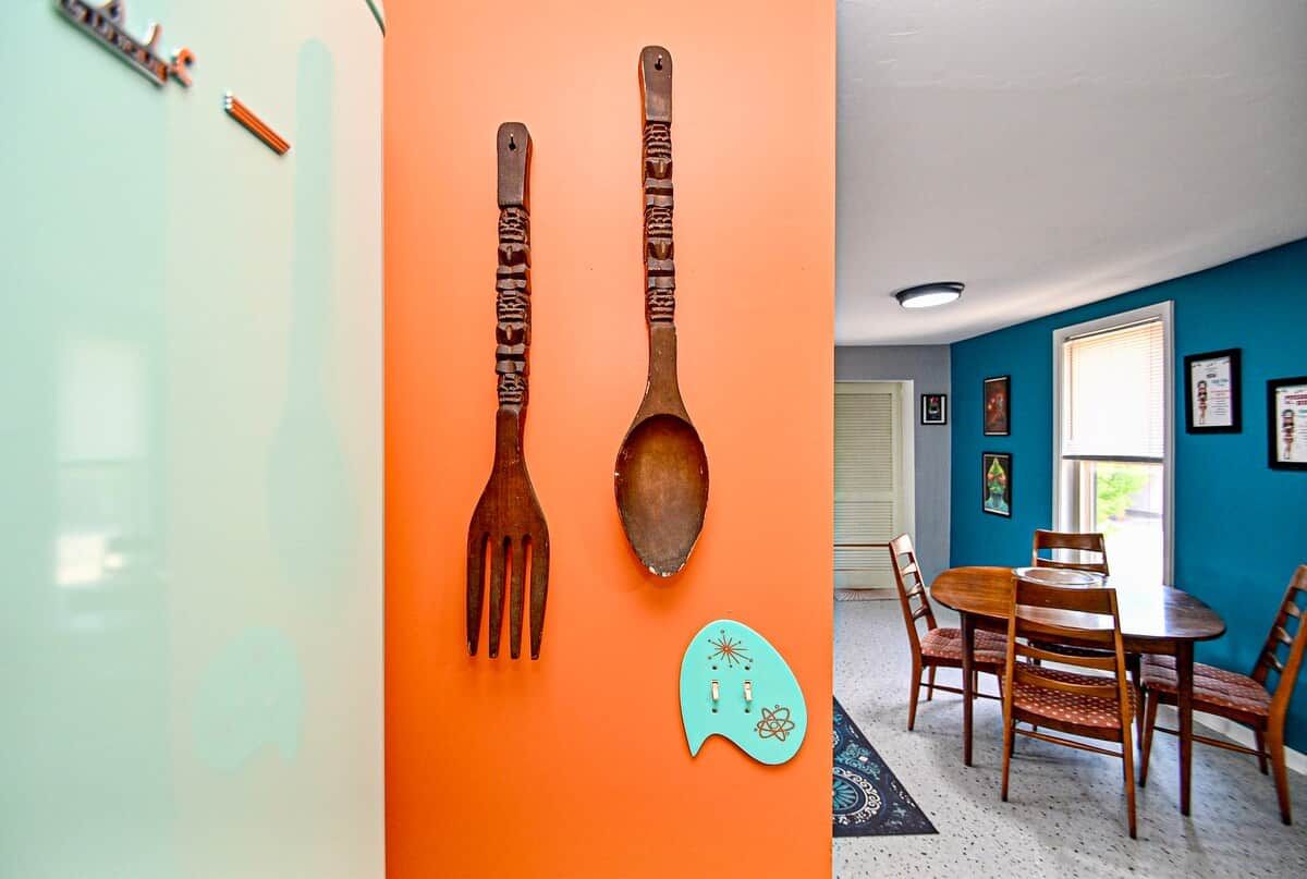 A room with a teal and orange wall, a wooden dining table with chairs, and a kitchen area with utensils mounted on the wall.