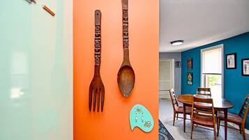 A room with a teal and orange wall, a wooden dining table with chairs, and a kitchen area with utensils mounted on the wall.