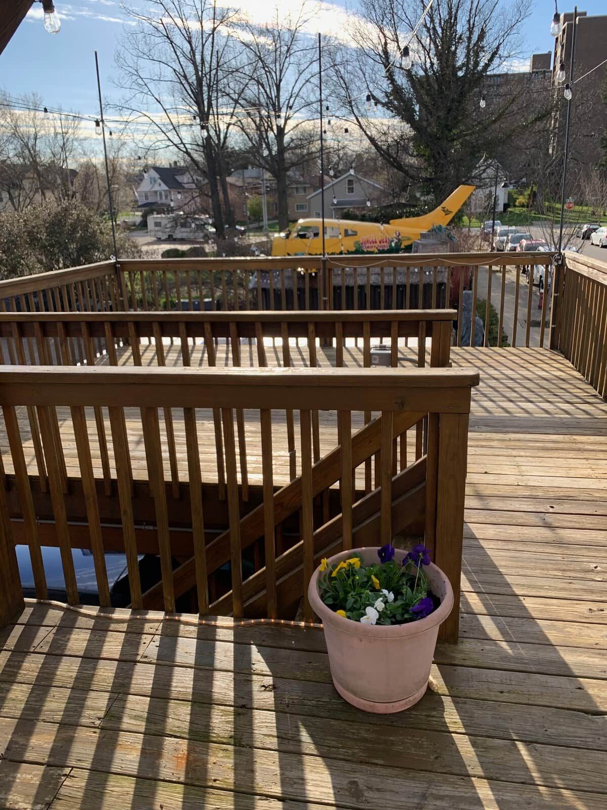 Une terrasse en bois avec une rambarde, un pot de fleurs et une vue sur une rue où l’on aperçoit un véhicule jaune.