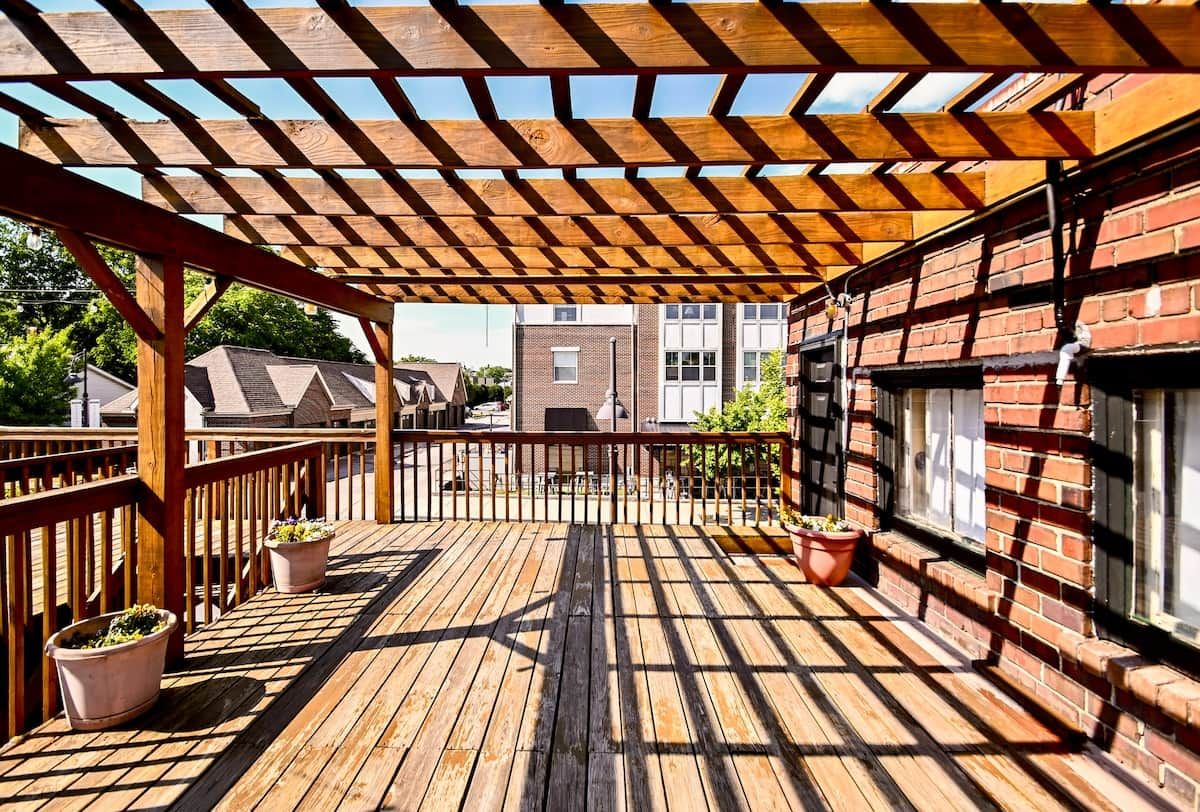 A wooden deck with a pergola, potted plants, and a brick building in the background.
