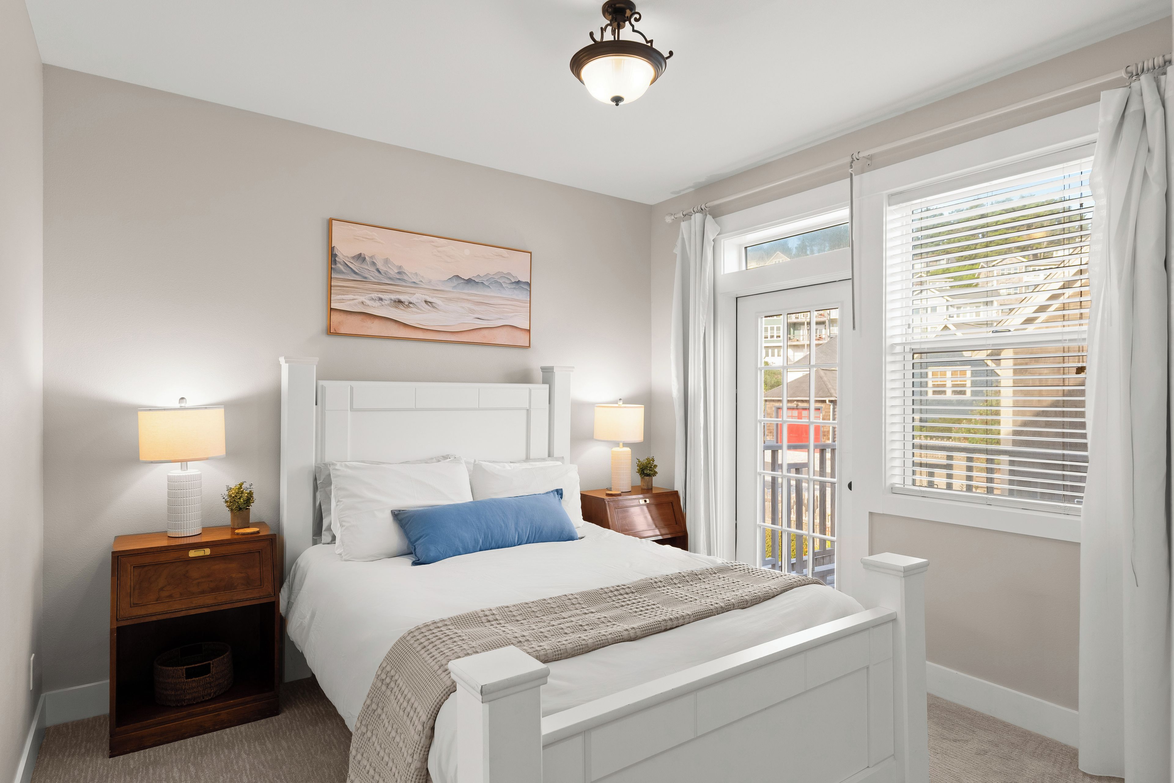 A bedroom with a bed, bedside lamps, a nightstand, a window with blinds, and a painting on the wall.