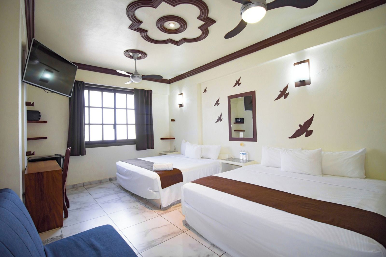 A hotel room with two beds, a desk, a chair, a TV, and a ceiling fan.