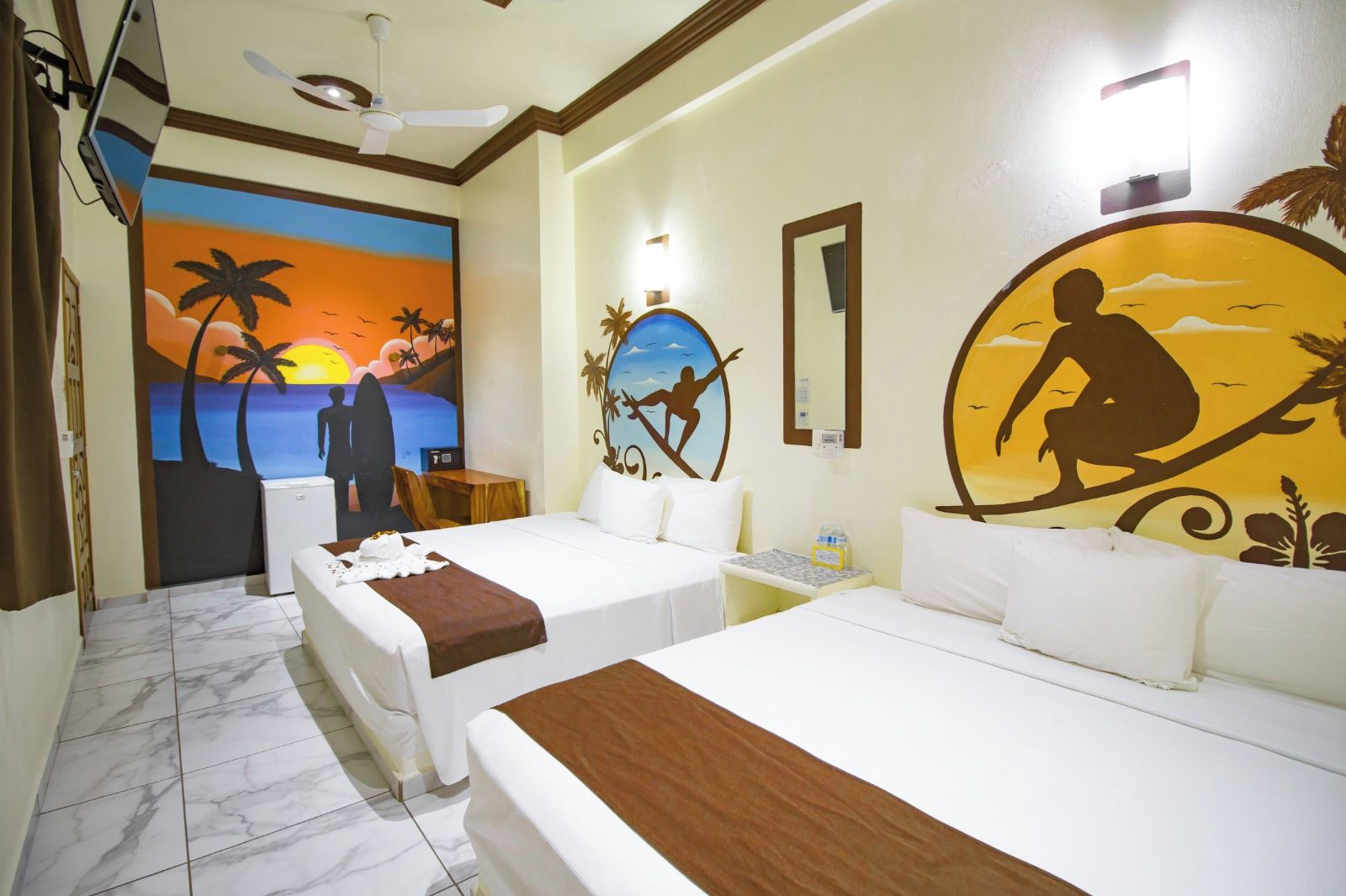 A hotel room with two beds, a ceiling fan, a painting of a sunset, and a wall-mounted mirror.