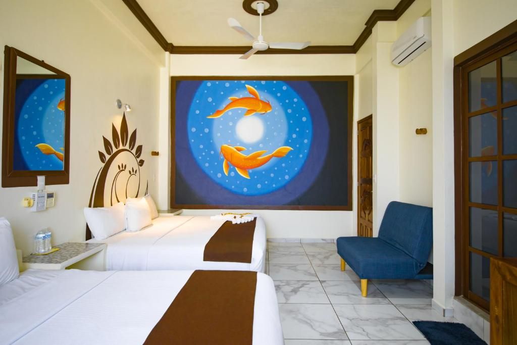 Habitación de hotel con dos camas, una silla azul, un ventilador de techo y un cuadro grande de peces en la pared.