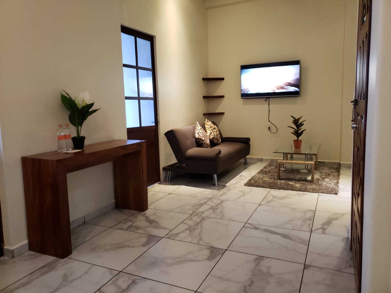 Una sala de estar con sofá, una mesa de centro, un televisor montado en la pared y una consola de madera con una planta y botellas de agua.