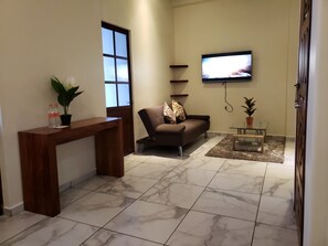 Una sala de estar con sofá, una mesa de centro, un televisor montado en la pared y una consola de madera con una planta y botellas de agua.