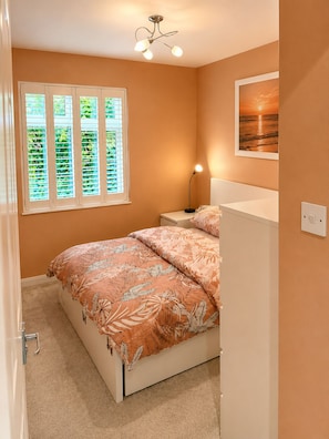 Ein Schlafzimmer mit einem Bett, einem Nachttisch, einer Lampe, einem Fenster mit Jalousien und einem gerahmten Bild an der Wand.