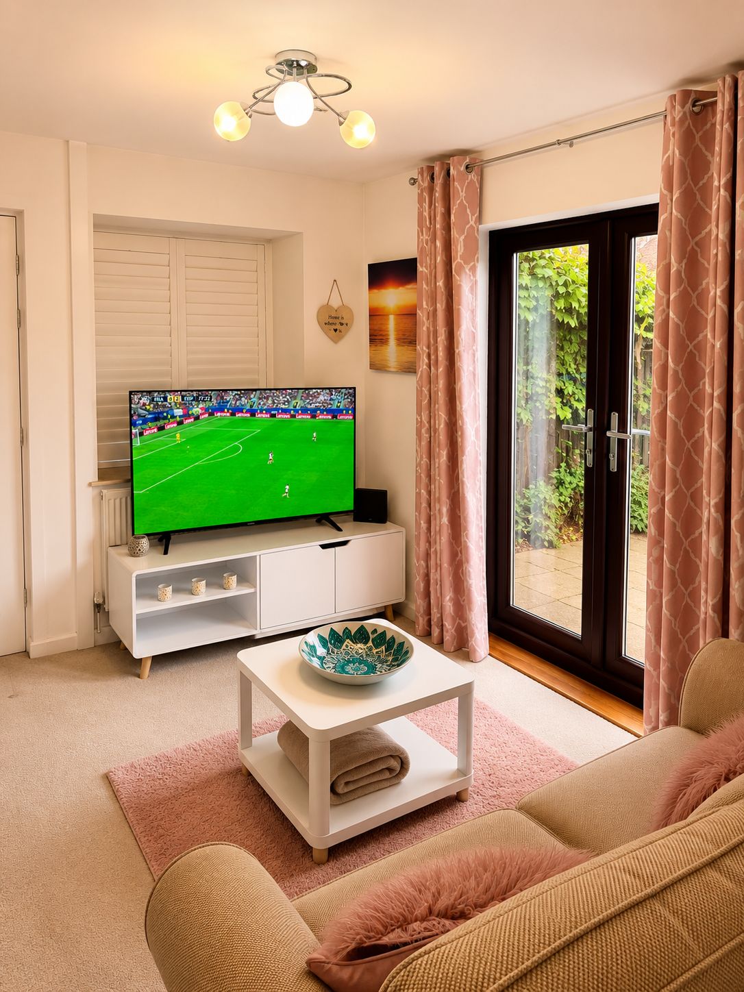 Ein Wohnzimmer mit Fernseher, der ein Sportevent zeigt, einem weißen Schrank, einer Schale auf einem Couchtisch und Blick auf Grün durch eine Glastür.