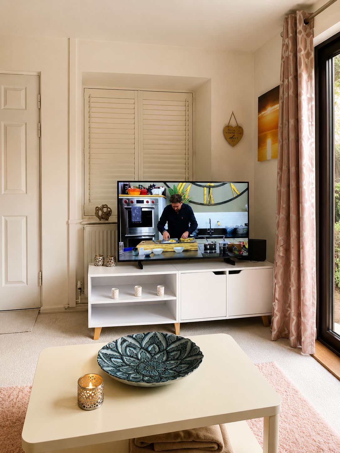 A modern living room with a television displaying a cooking show, a white cabinet with kitchenware, a decorative plate, and a window with curtains.