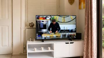 A modern living room with a television displaying a cooking show, a white cabinet with kitchenware, a decorative plate, and a window with curtains.