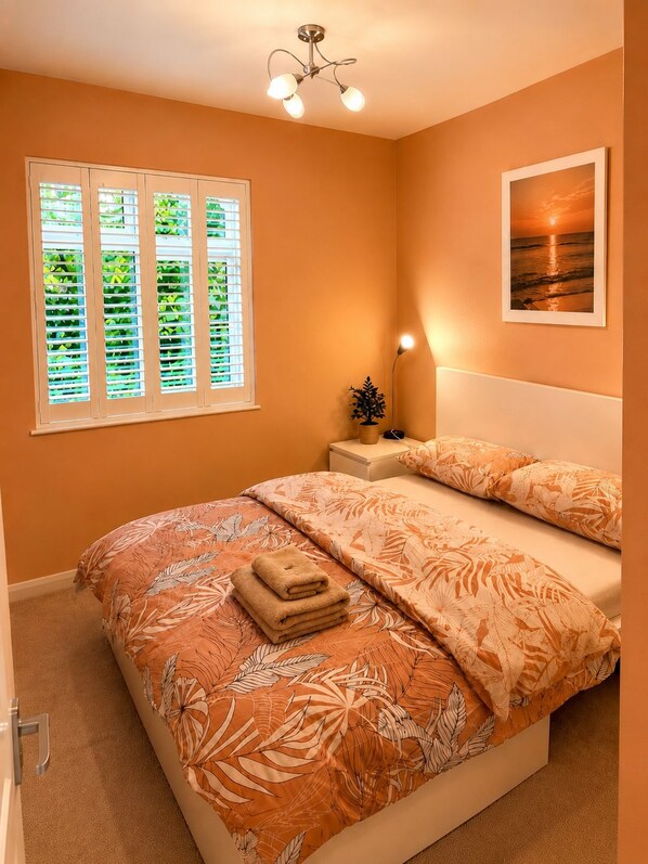 Ein Schlafzimmer mit einem großen Bett, einem Fenster mit Jalousien, einem Nachttisch mit Lampe und einem gerahmten Bild an der Wand.