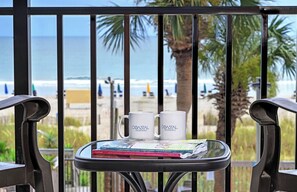 A balcony with two white mugs labeled 'Coastal Real Estate' and a magazine on a glass table, overlooking a beach with palm trees and the ocean. - 1BR Oceanfront Suite, Wake Up to the Atlantic! (Myrtly Beach)