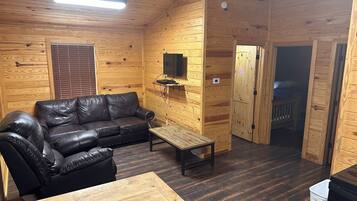 Interno di una baita in legno, con un divano in pelle, un tavolino, una televisione montata a parete e una porta che dà su un'altra stanza.