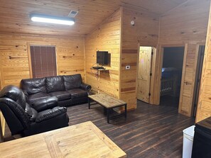Interior de uma cabana de madeira com um sofá de couro, uma mesinha, uma televisão fixada na parede e uma porta que dá para outro quarto.