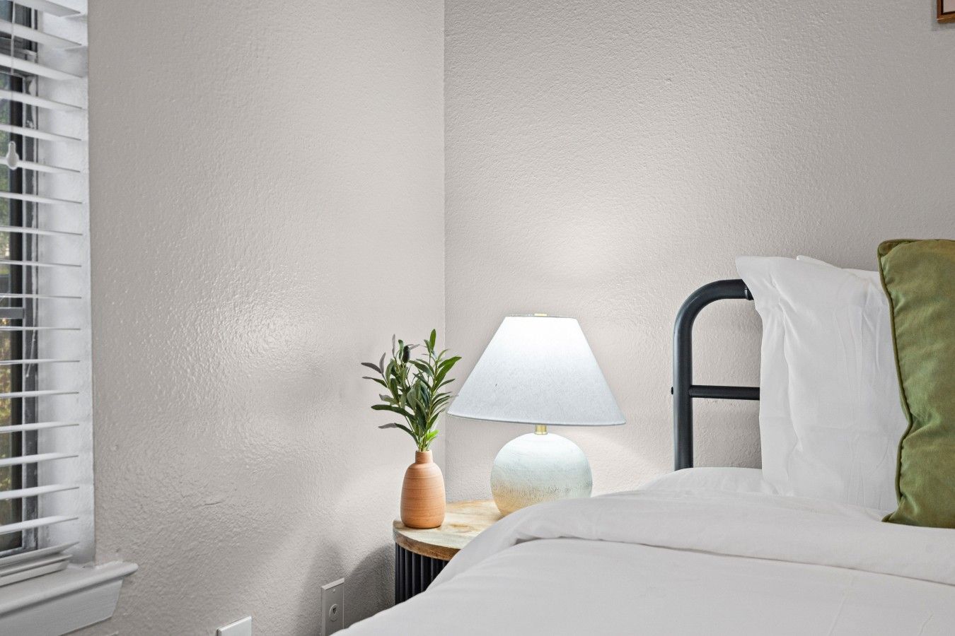 A bedroom with a bed, a bedside table with a lamp, and a window with blinds.