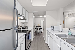 Una cocina moderna con armarios blancos, horno empotrado, microondas y pileta. Se ve un área de comedor con una mesa y sillas al fondo.